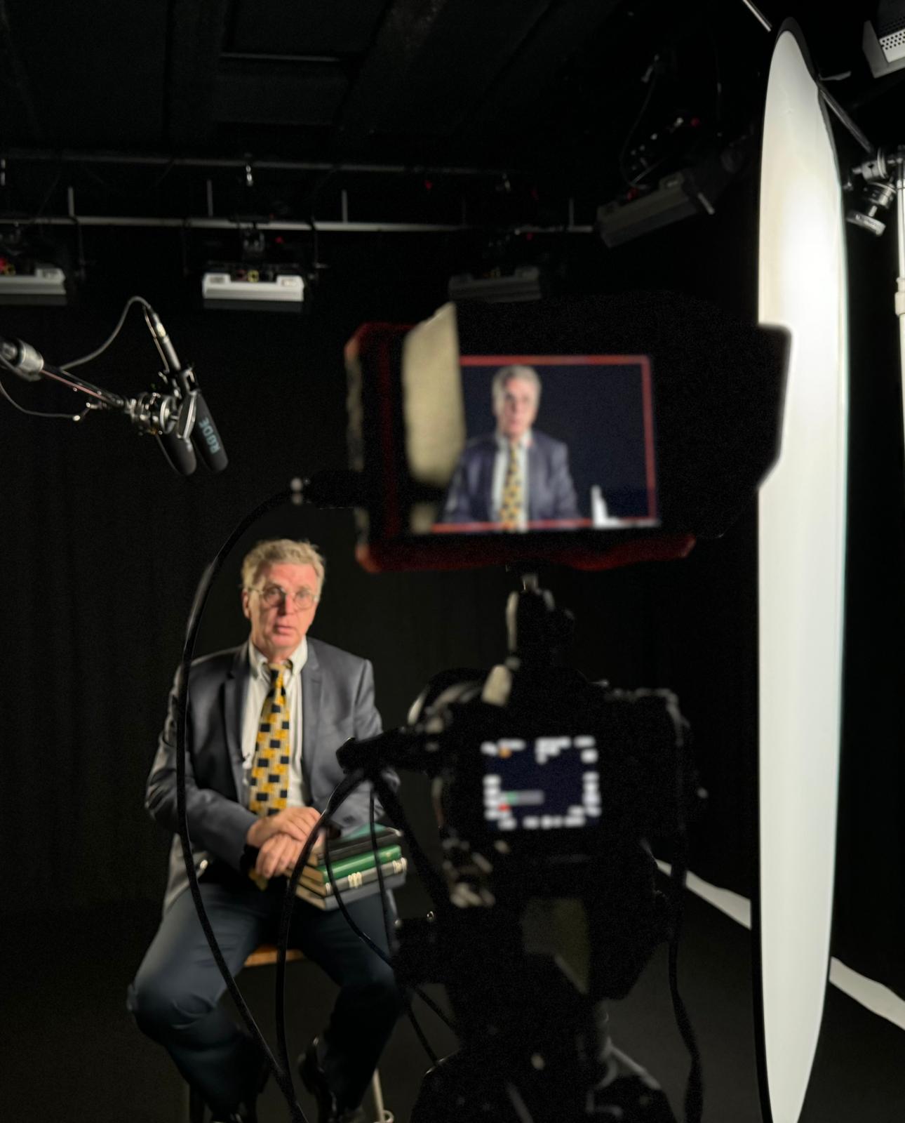 A man seated for a filmed interview behind the camera and studio lighting